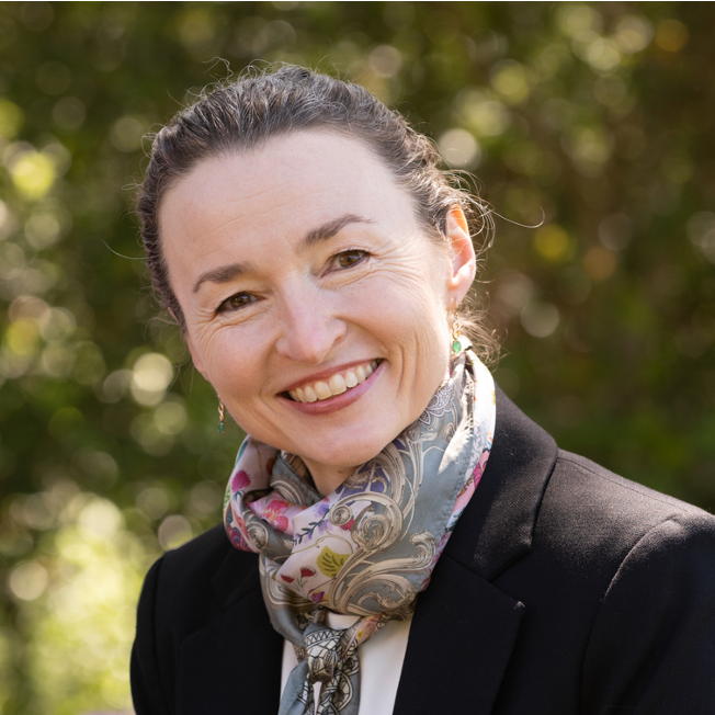 A headshot of Joanna Gardner smiling with a background of blurry green trees.
